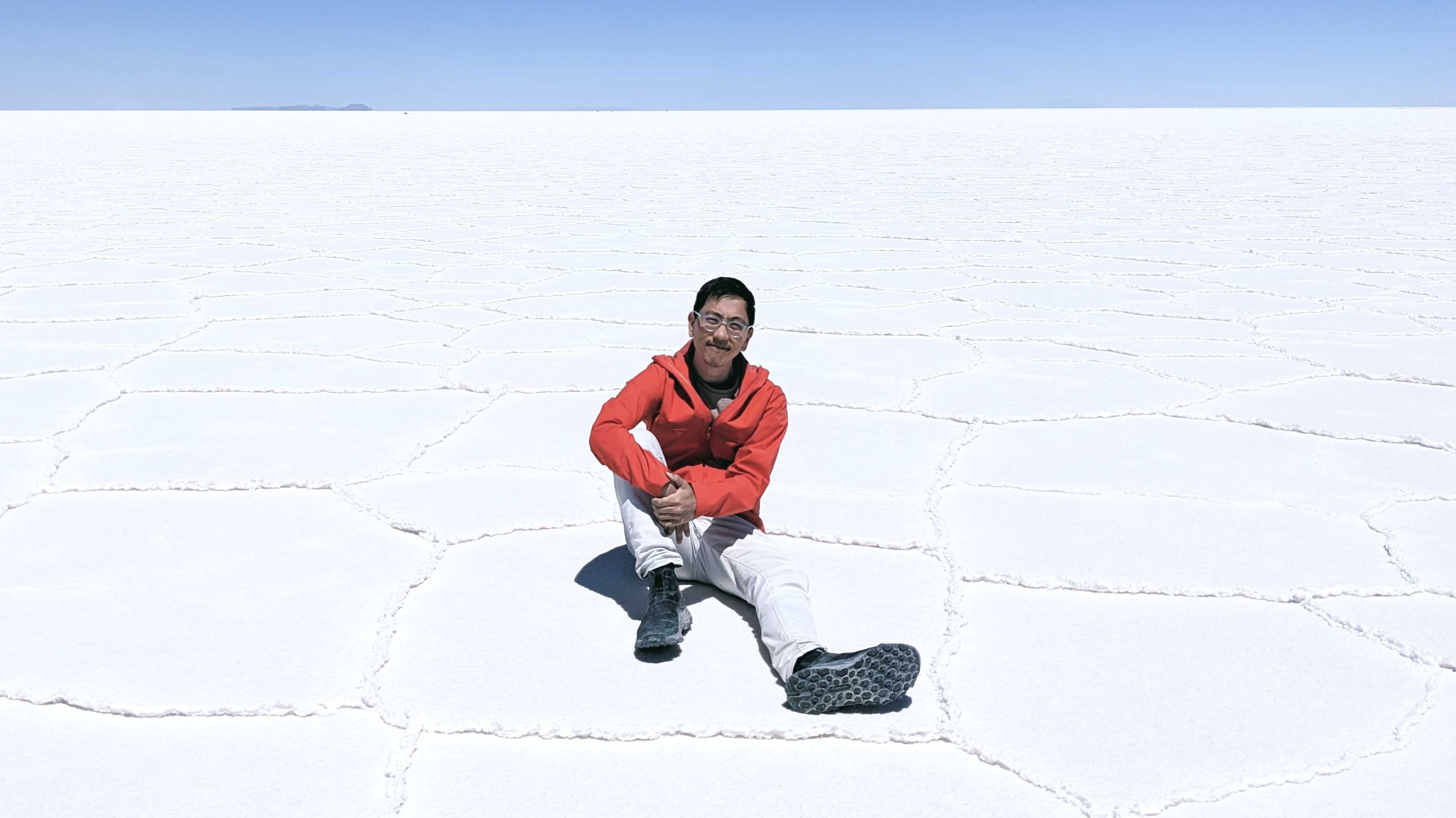 Henry in a red coat and white jeans sitting on the ground of the Uyuni Salt Flats in Bolivia, a barren stark white landscape against a clear blue sky.